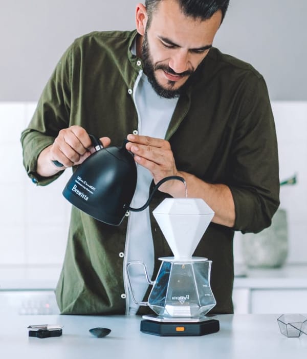 Barista pouring water from a black Brewista kettle into a white diamond shaped Brewista brewer balancing on a glass coffee pot.